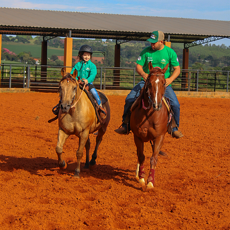 EMPRESA-08 Grupo de pessoas vestindo camisas verdes e chapéus de cowboy posando ao lado de um cavalo em um evento da ABQM (Associação Brasileira de Criadores de Cavalo Quarto de Milha). Na frente, uma mesa com diversos troféus e arranjos de flores. O fundo exibe um painel com os logos e nomes das competições '17ª Copa dos Campeões', '45º Potro do Futuro' e '10º Derby'.
