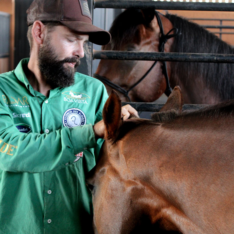 Treinador Marquinhos Um homem sorridente usando um chapéu de cowboy, segurando a cabeçada de um cavalo preto. O ambiente é ao ar livre, com uma estrutura coberta ao fundo e vegetação visível.